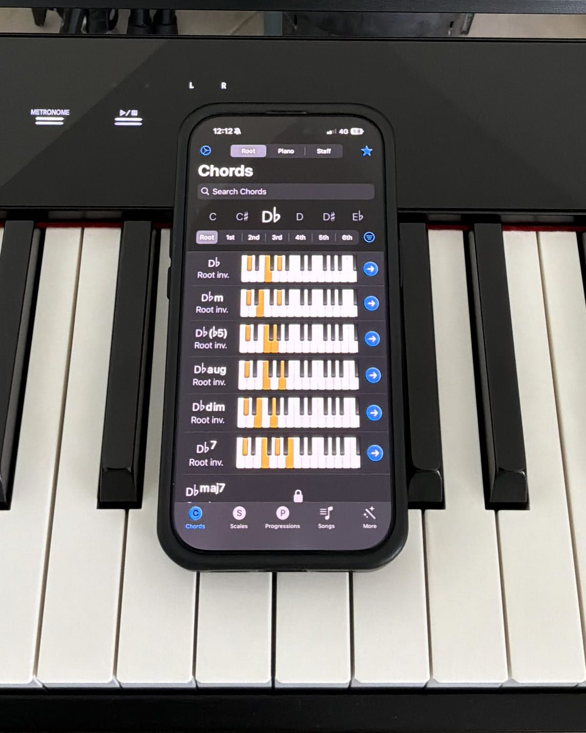 The Piano Chords and Scales app open on an iPhone resting on a digital piano, listing D-flat major, minor, flat-five, augmented, diminished and seventh chords, each drawn on a small keyboard with the chord tones highlighted.