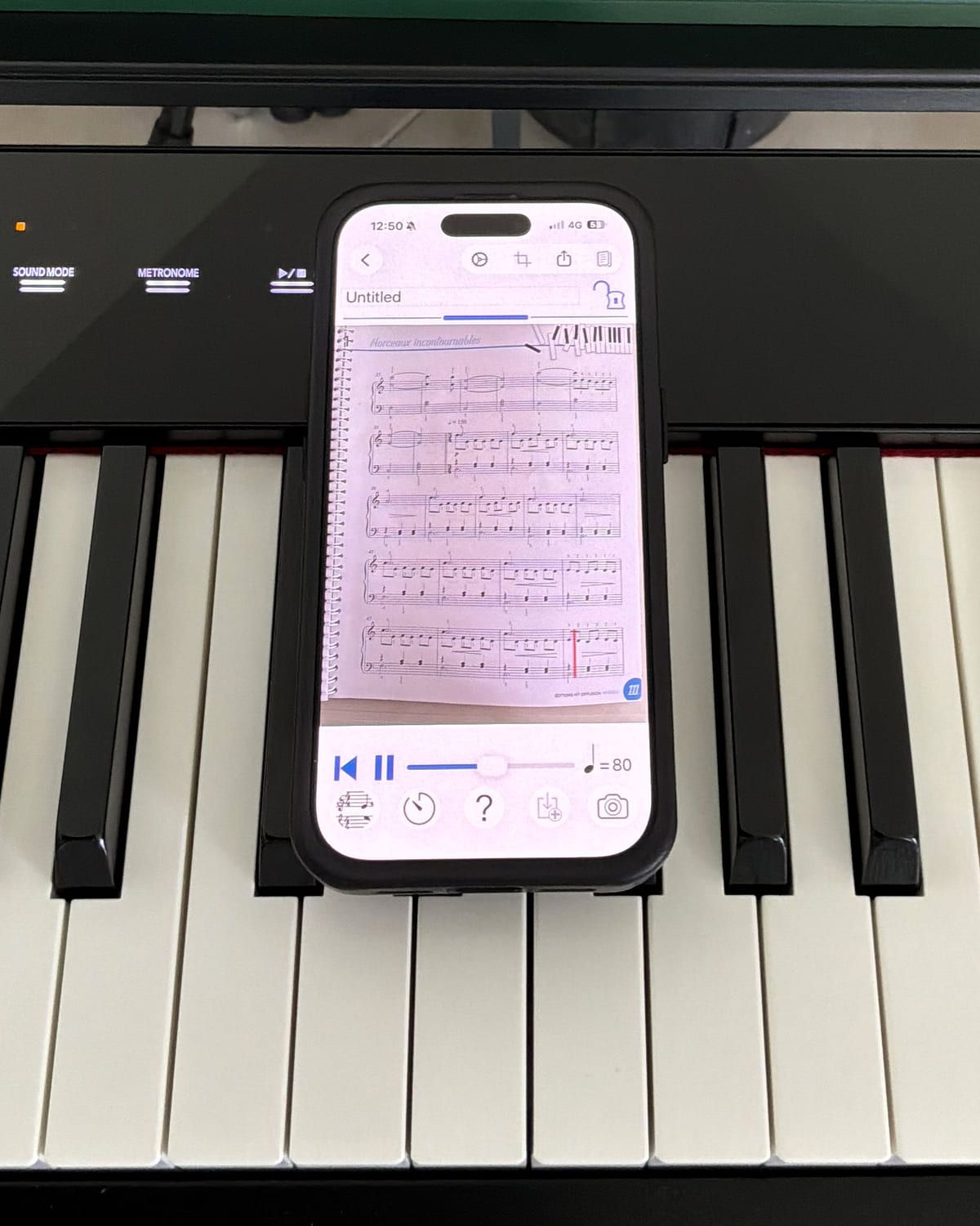 PlayScore 2 on an iPhone propped on a digital piano, playing back a photographed page of piano sheet music at 80 bpm with a red playback cursor part-way down the last system.